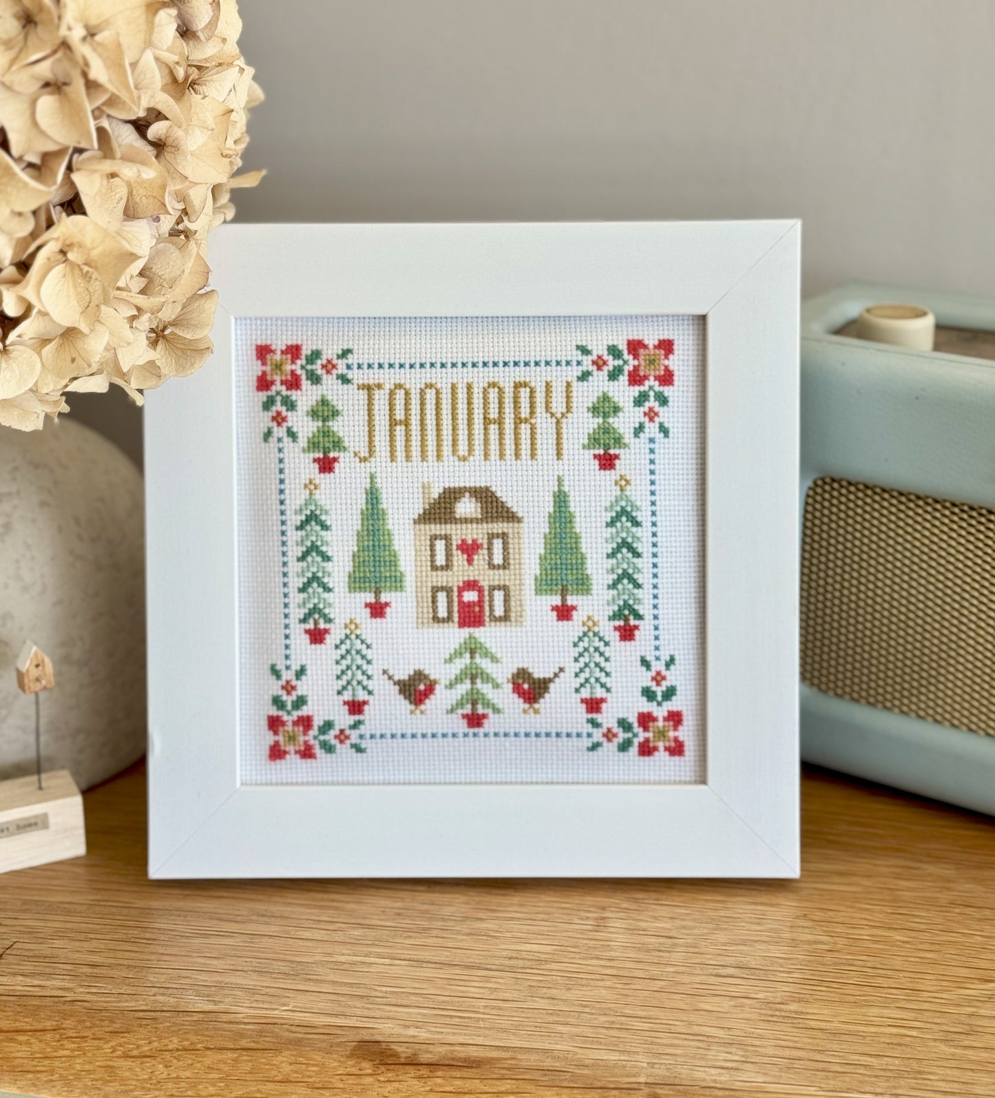 Framed cross-stitched artwork with 'January' text and festive design on a wooden surface.